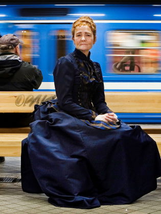 Ann Petrén i en gammeldags klänning sittandes på en bänk på en tunnelbaneperrong
