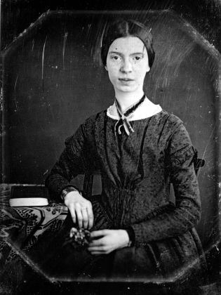 Ung kvinna i 1800-talsklänning sitter vid ett bord med en bok, ser in i kameran i svartvitt porträtt.