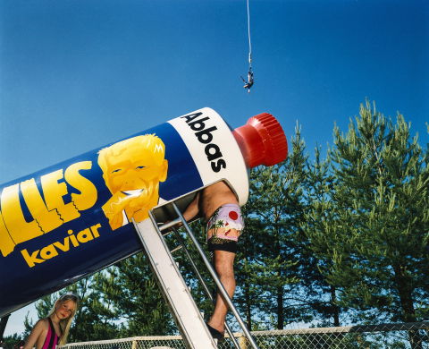 Man i badshorts klättrar in i en jättetub med Kalles kaviar-design; kvinna står bredvid, person bungyhoppar i bakgrunden.