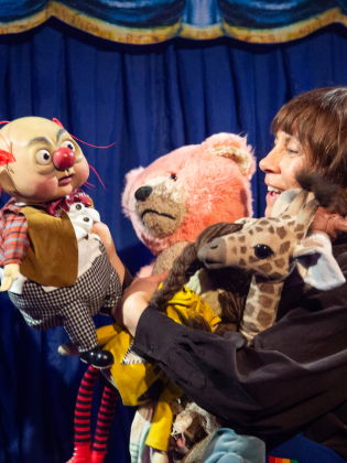 Kvinna håller flera dockor framför blå ridå, inklusive clown, giraff och rosa björn.