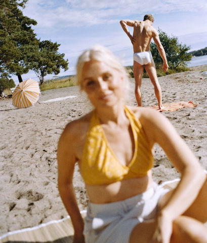Kvinna i gult bikinilinne sitter suddigt i förgrunden på en sandstrand, medan en man i vita badkläder står i bakgrunden vid vattnet.