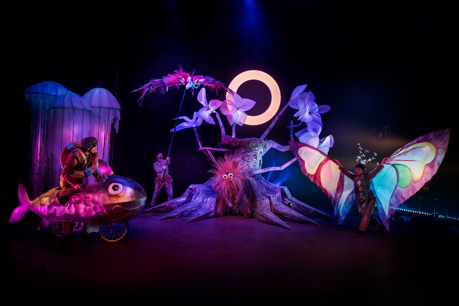 En skådespelare med fjärilsvingar, en med en cykel i form av en fisk, mitt i allt står ett träd. 