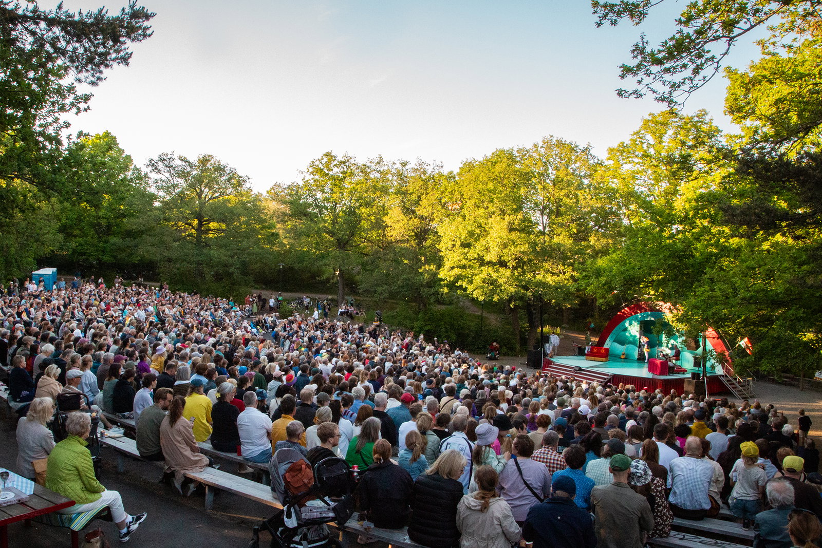 En stor publikgrupp som sitter framför en utomhusscen där det spelas teater en somrig kväll.