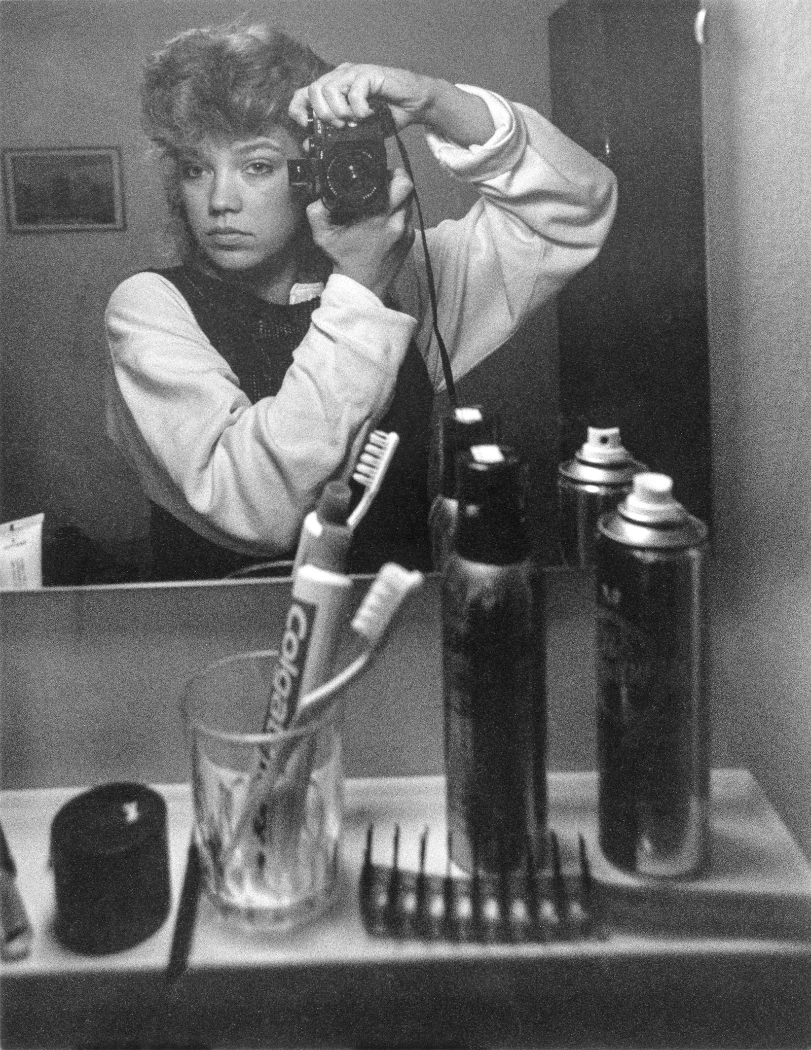 Ung kvinna med kort lockigt hår tar spegelselfie med kamera i badrum; tandborste, tandkräm och sprayburkar på handfat.