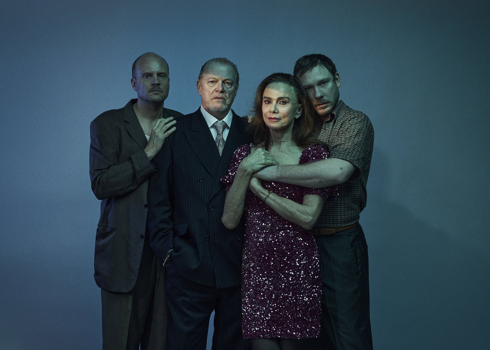 Fyra vuxna står tätt: skallig man i mörk kavaj, äldre man i kostym, kvinna 50–60 i röd paljettklänning, ung man håller om henne.