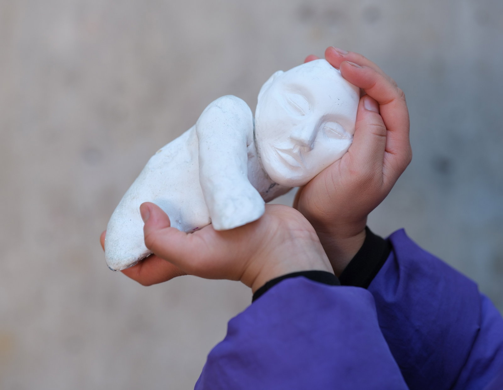Ett person håller en liten vit skulptur av en liggande figur med slutna ögon i sina händer. Närbild mot en mjuk, neutral bakgrund.