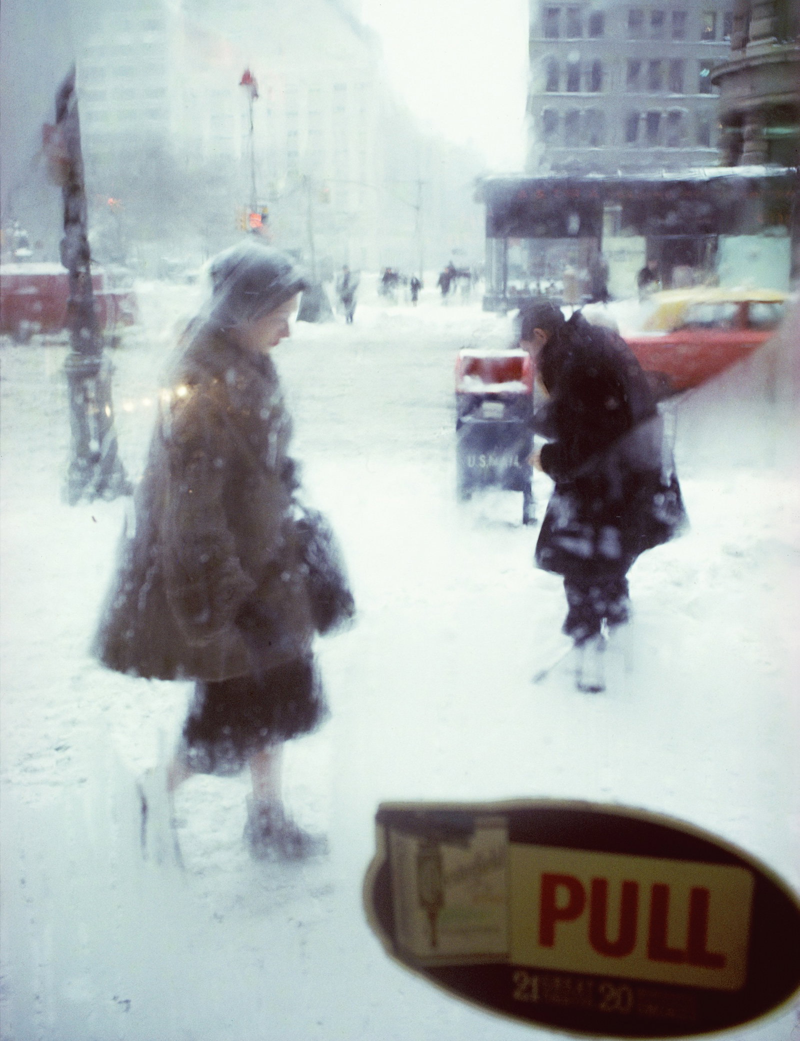 Två personer går genom ett snöigt stadslandskap i kraftigt snöfall, fotograferade genom en glasdörr med skylten ”PULL”.