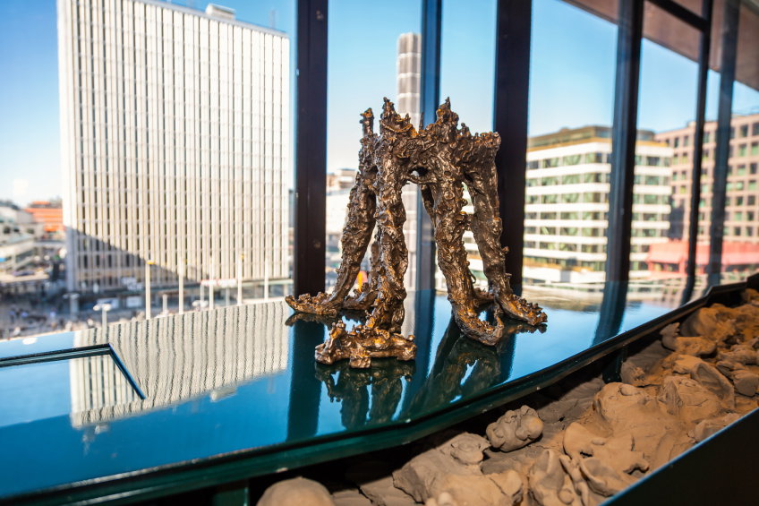 Bronsliknande skulptur med flera benliknande pelare på blank yta, utsikt över höghus i bakgrunden.