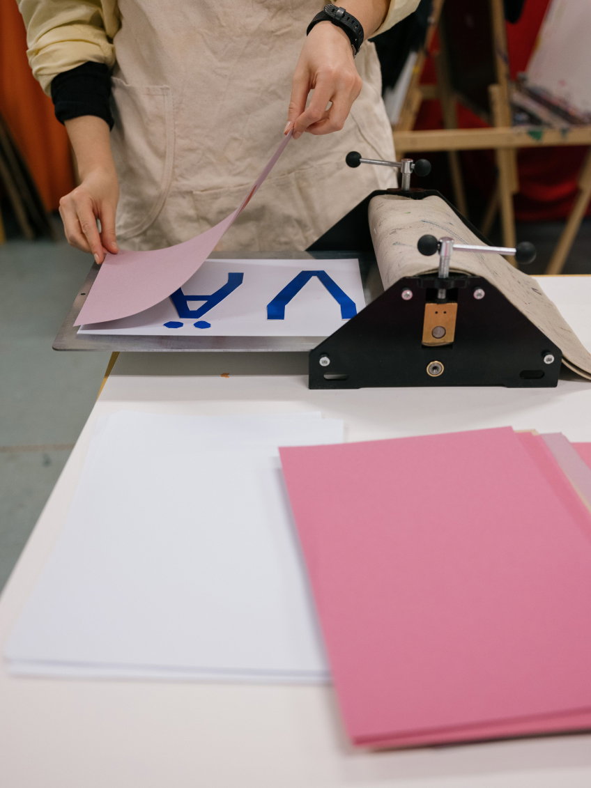 Person i beige förkläde placerar rosa papper på tryckpress.