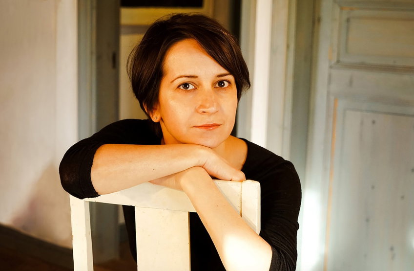 Kvinna ca 30–40 år med kort mörkt hår och svart långärmad tröja, lutar armarna på vit stol inomhus och ser mot kameran.
