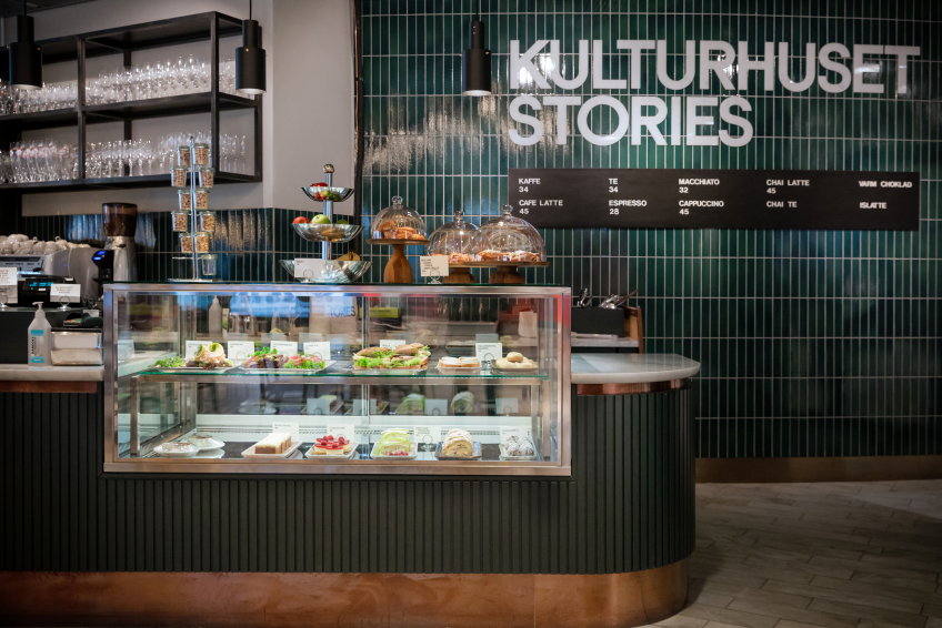 Cafédisk med smörgåsar och bakverk i glasmonter. Grön kakelvägg bakom med texten “Kulturhuset Stories” och meny.