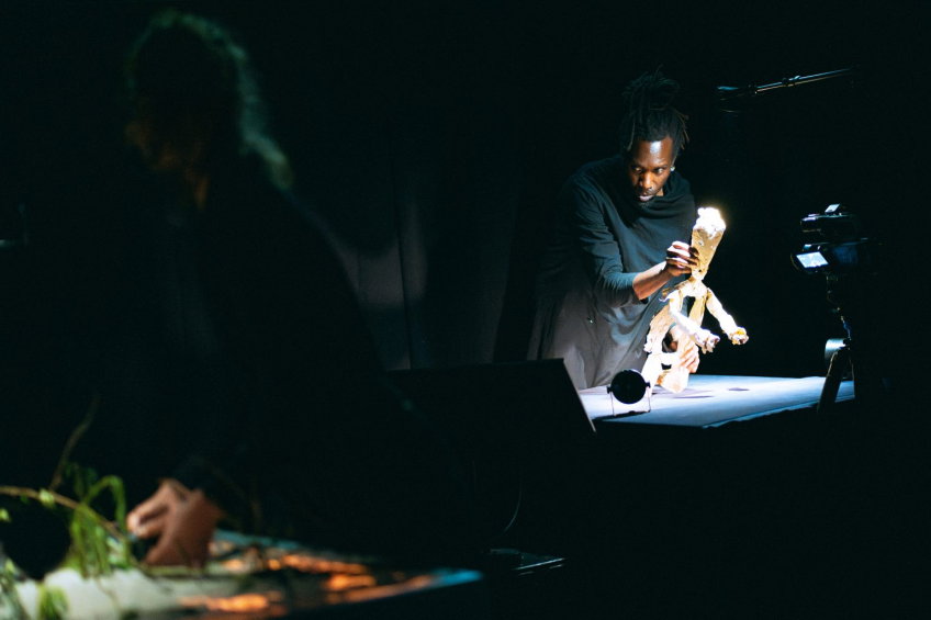 En person styr en trädocka på en mörk scen, upplyst av ett riktat ljus medan en annan person arbetar i bakgrunden.