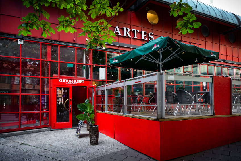 Uteservering utanför röd byggnad med skylten “Artes”. Bord och stolar under gröna parasoller vid entrén till Kulturhuset.