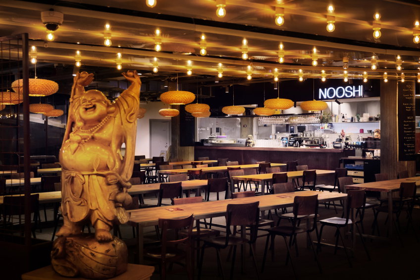 Restaurang med långbord och stolar, varm belysning i taket och en Buddhafigur i förgrunden. Baren “Nooshi” syns i bakgrunden.