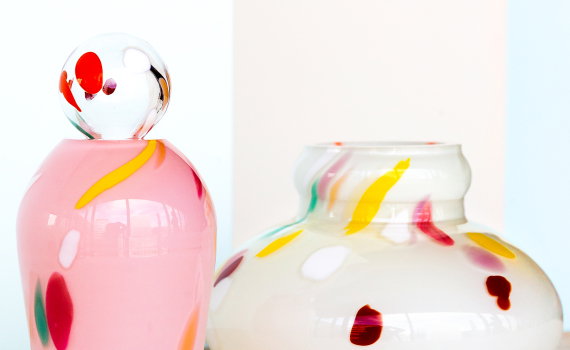Två färgglada glasföremål. En rosa vas med lock pryds av lekfulla färgfläckar, bredvid en vit skål med spridda, glada penseldrag i olika nyanser.