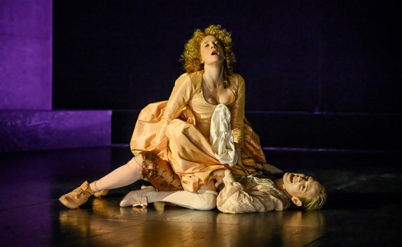 Två personer i 1700-talskläder på en scen, den ena sitter över den andra som ligger på golvet.