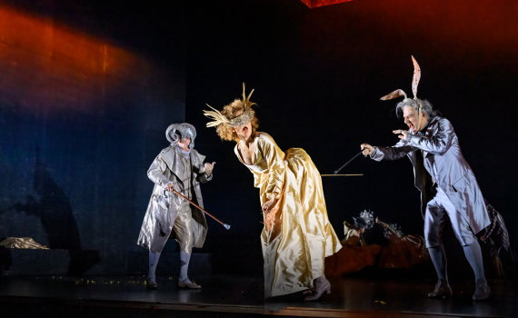 Tre skådespelare i barockkostymer, en i gul klänning med mask, omgiven av två i djurinspirerade dräkter på en mörk scen.