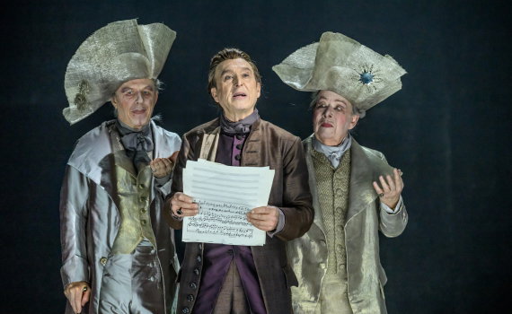 Tre män i 1700-talskläder, en håller noter medan de andra två bär stora silverfärgade hattar.