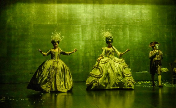 Två kvinnor i gröna 1700-talsklänningar står med utsträckta armar, en man i bakgrunden med mask.