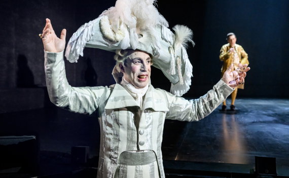 Skådespelare i vit barockkostym och stor fjäderhatt gestikulerar, medan en man i gulddräkt syns i bakgrunden.