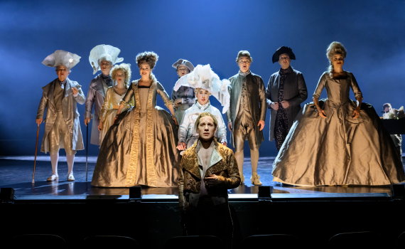 Grupp på nio personer i 1700-talskläder står upplysta på scen, en man i guldbroderad jacka längst fram.