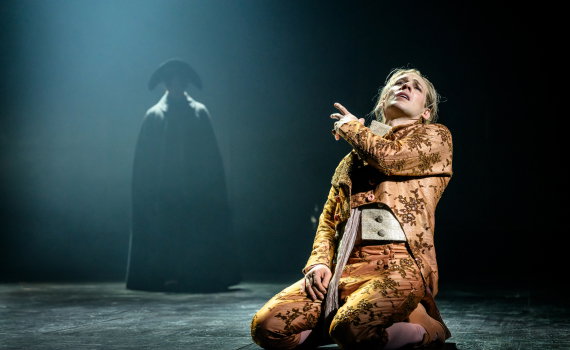 Man i guldbroderad rock knäböjer och gestikulerar, en mörkklädd figur står i bakgrunden i dimma.