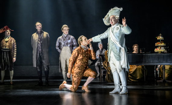 Man i vit 1700-talsdräkt låter en annan person i orange kläder kyssa hans hand, fler personer i bakgrunden.