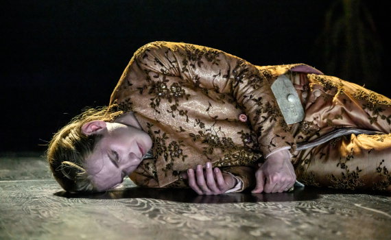 Man i guldbroderad dräkt ligger på golvet med huvudet på sidan, blicken riktad mot kameran.