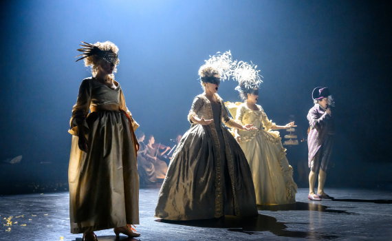 Fyra personer i 1700-talskläder på scen, tre kvinnor i stora klänningar och en man med hatt, ljus bakgrund.