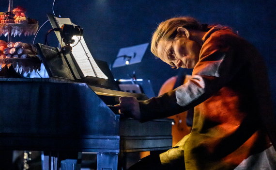 Person i 1700-talskläder sitter vid ett piano med noter, huvudet lutat mot tangenterna.