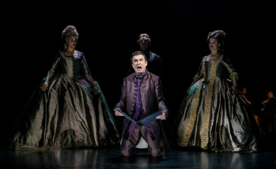 Man i mörk dräkt knäböjer med papper i händerna, två kvinnor i stora klänningar bakom.