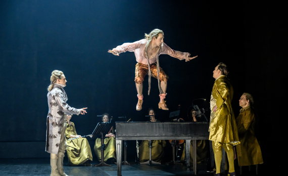 Person i orange byxor hoppar upp på ett piano, tre andra personer ser på.