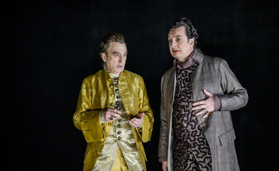 Två män i 1700-talskläder, en i gul dräkt och en i mönstrad rock, ser bekymrade ut.