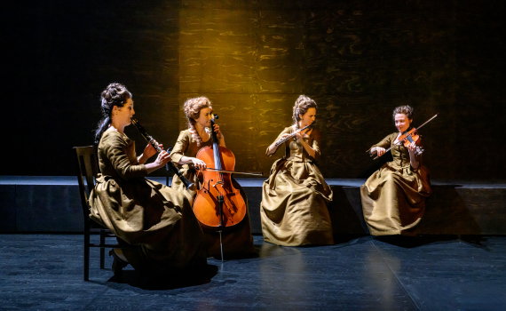 Fyra kvinnor i bruna 1700-talsklänningar spelar blåsinstrument, cello och violin.