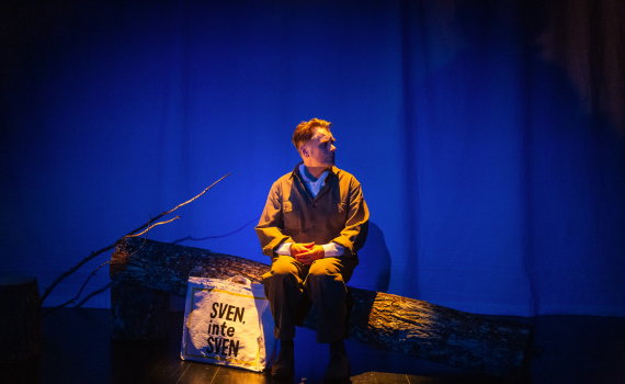 Sven Björklund sitter på en stock i ett blått ljussatt rum med en kasse bredvid sig som det står Sven, inte Sven på.