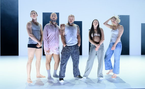 Fem vuxna i smutsiga kläder står på rad på scen med ljus bakgrund och mörka rektanglar bakom sig.