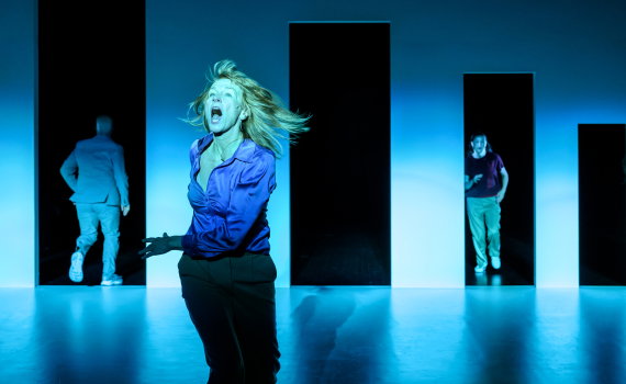 Kvinna i lila blus skriker i förgrunden, två män syns i bakgrunden mellan höga, svarta dörrliknande öppningar.