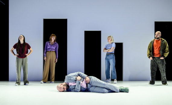 Fem personer står på scen, en man i kostym ligger ner medan de andra tittar åt olika håll.