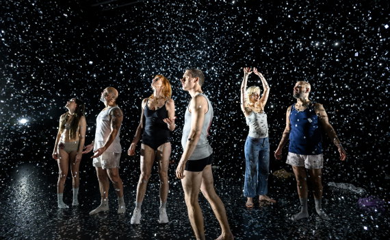 Sex vuxna i underkläder och linnen står under fallande konfetti på en mörk scen.