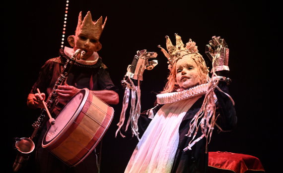 Två vuxna personer i masker och kronor står på scen; en spelar trumma och blåsinstrument, den andra håller rekvisita.