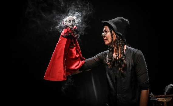 Vuxen kvinna i hatt och glasögon håller en liten docka i röd mantel på en mörk teaterscen.