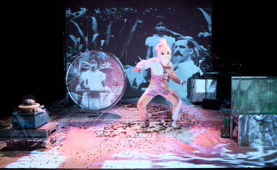 Person i vit overall och vit mask står på scen med böjd kropp, projektion och teknisk utrustning bakom.