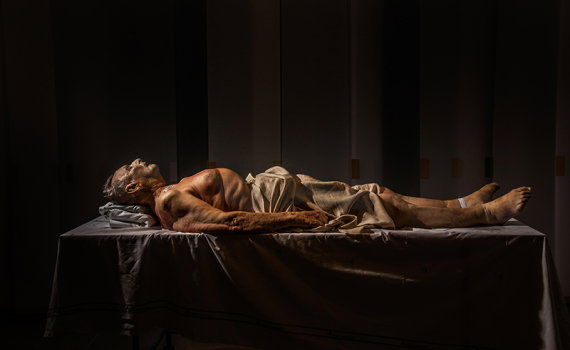 Äldre man med grått hår ligger naken på brits med lakan över höfterna, armband på handleden, mörk bakgrund.