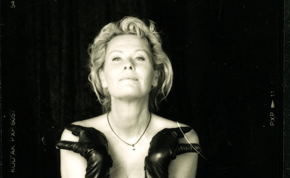 Medelålders kvinna med kort blont hår och svarta handskar poserar barbröstad mot mörk bakgrund.