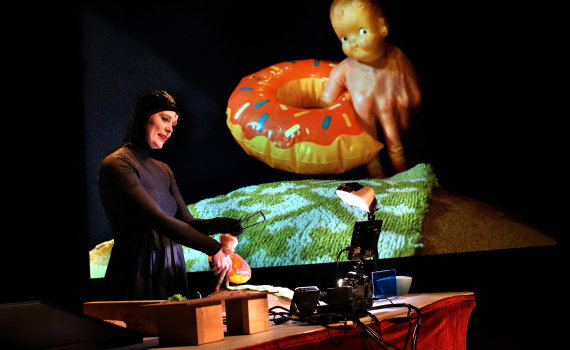 Kvinna i mörk kroppsnära dräkt står vid bord med kamera och lampor, hanterar en docka framför projicerad bild.