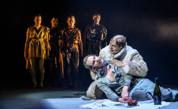 Två män ca 30–40 år i slitna kläder sitter på golv; en håller den andre. Fyra personer i blanka dräkter står bakom.