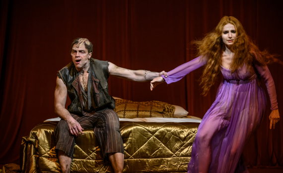 Man ca 30 år i smutsig väst sitter på säng och håller kvinna ca 30 år i lila klänning framför röd ridå.