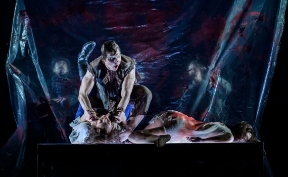 Man ca 30 år i väst sitter grensle över kvinna i vit klänning på bord, plastskynke med röda fläckar bakom.