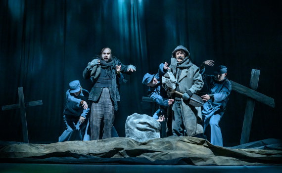 Två män ca 40–60 år i rockar står vid gravkors, tre personer i blå uniformer omger dem på scen.