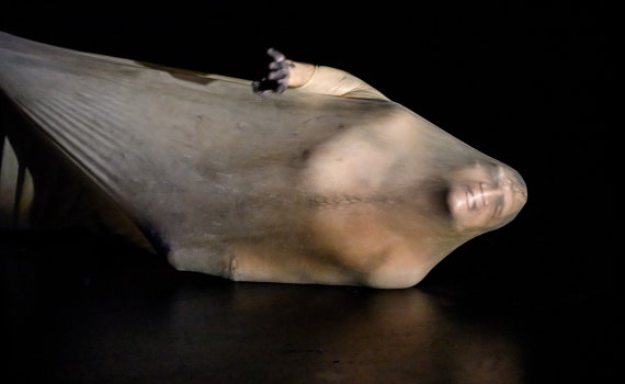 Halvnaken man ca 30 år ligger insvept i genomskinlig plast på mörkt golv, ena armen sträckt upp.